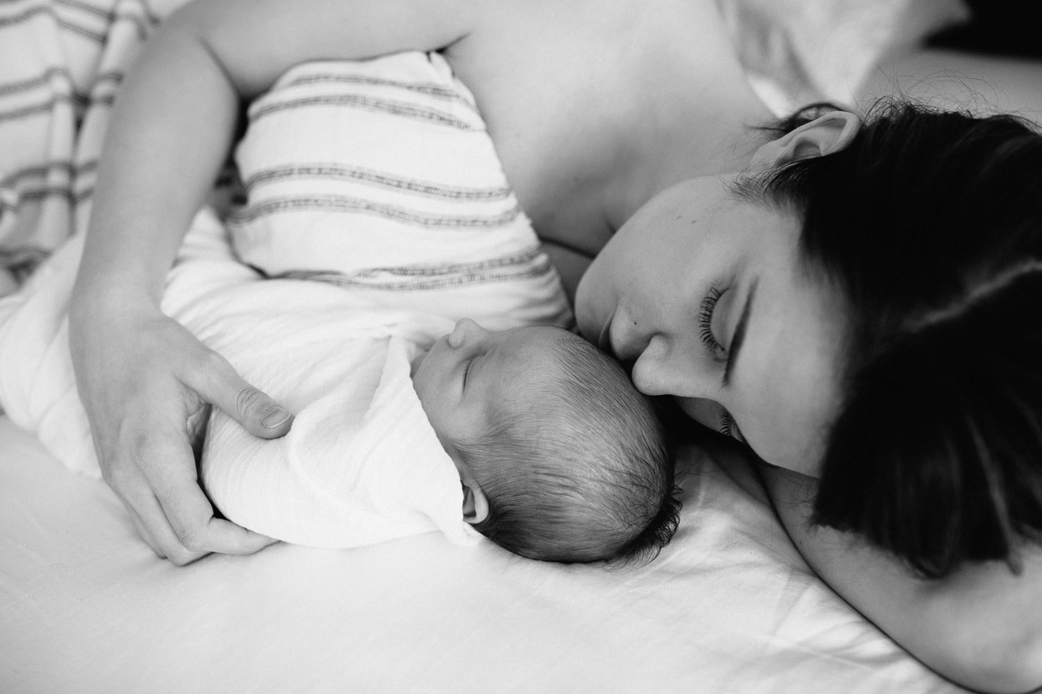 new mother with long dark hair lying on bed next to sleeping 2 week old baby boy wrapped in white swaddle, kissing son on head - Markham Lifestyle Photos