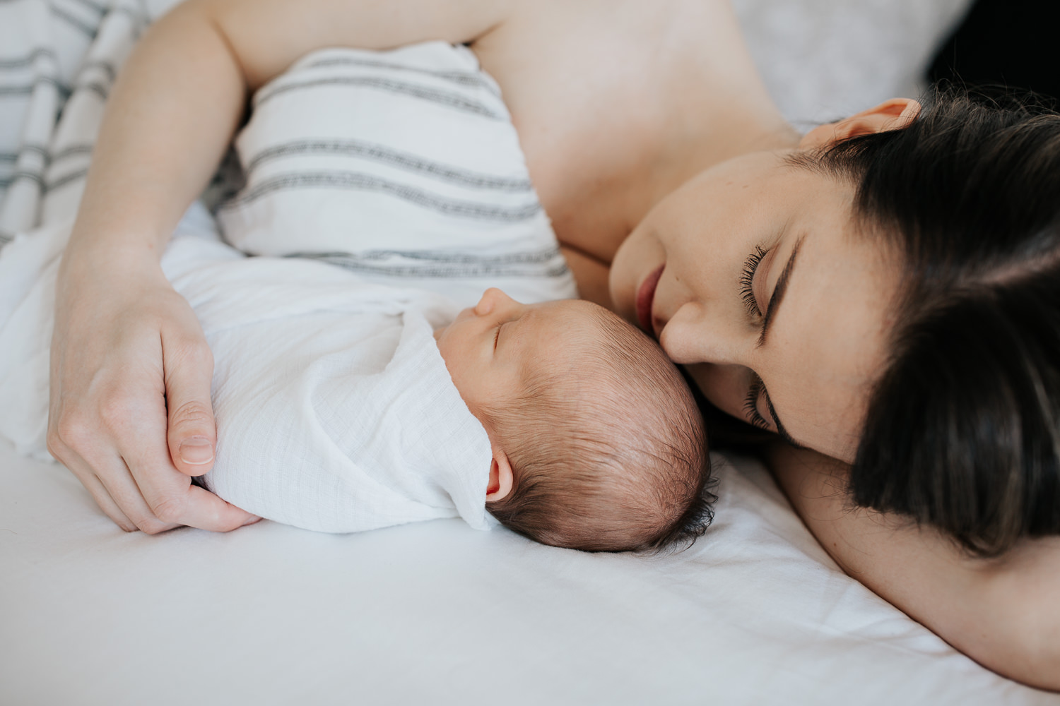 new mother with long dark hair lying on bed next to sleeping 2 week old baby boy wrapped in white swaddle - Barrie Lifestyle Photos