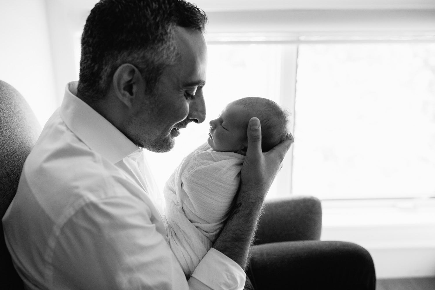 new dad sitting in nursery rocking chair, holding sleeping 2 week old baby boy to his face, smiling at son - GTA Lifestyle Photos