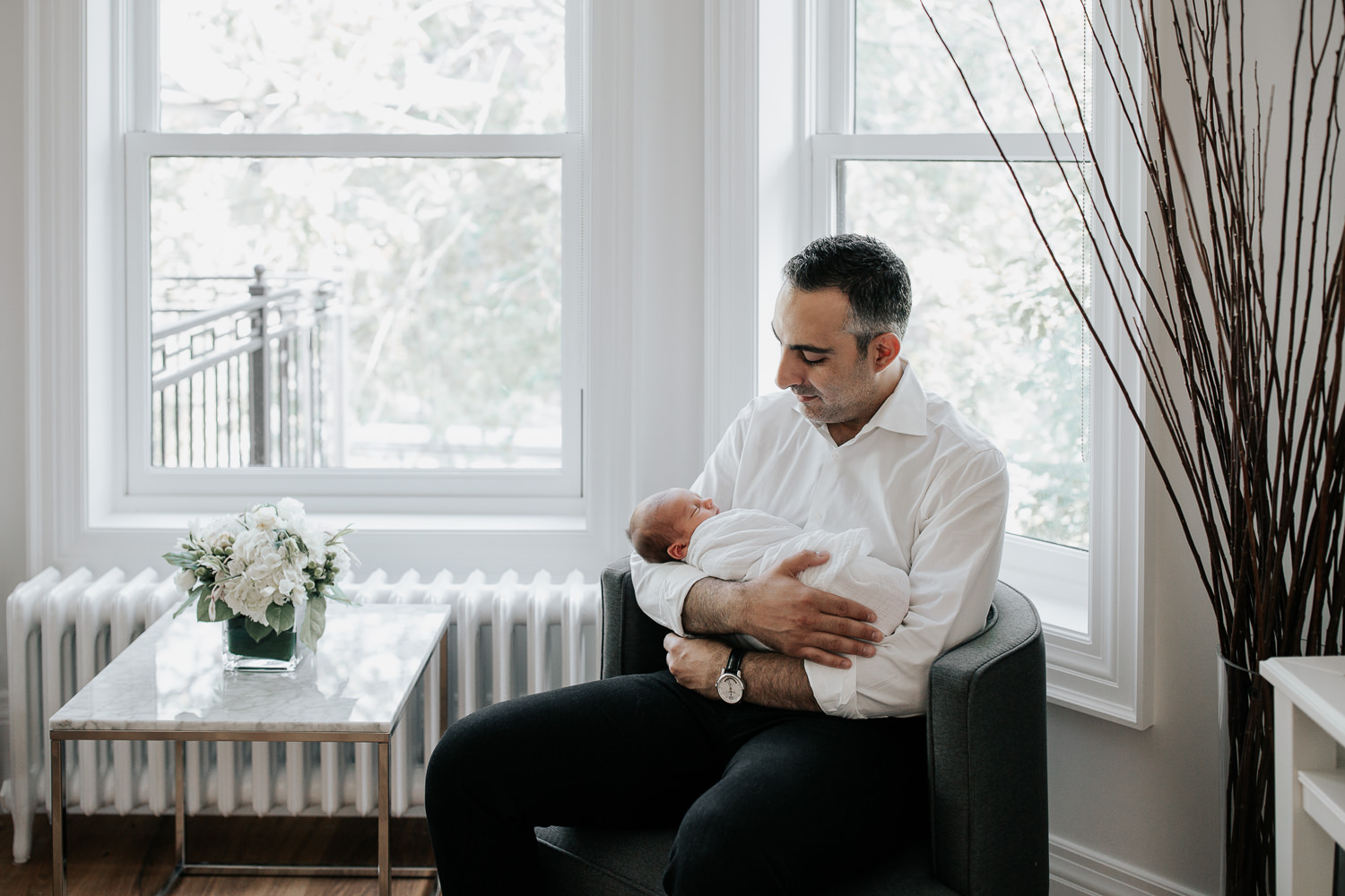 new father sitting in chair in living room, holding sleeping, swaddled 2 week old baby boy in his arms, smiling at son - Markham Lifestyle Photography