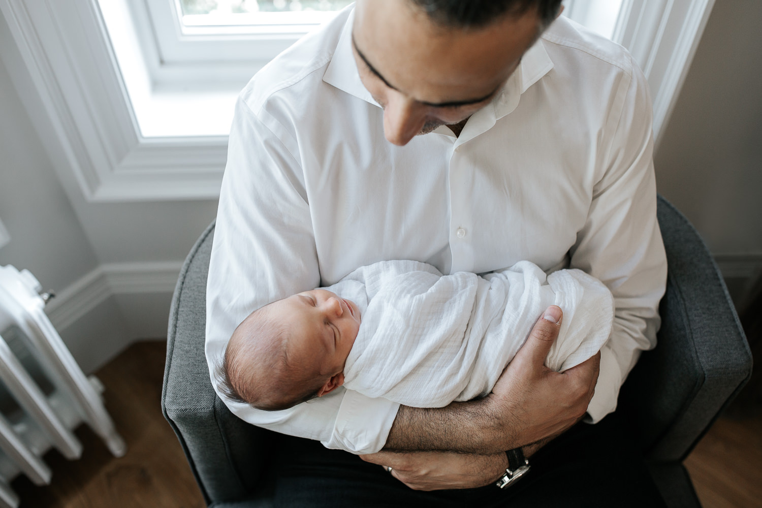 new father sitting in chair in living room, holding sleeping, swaddled 2 week old baby boy in his arms, looking at son - Barrie Lifestyle Photography