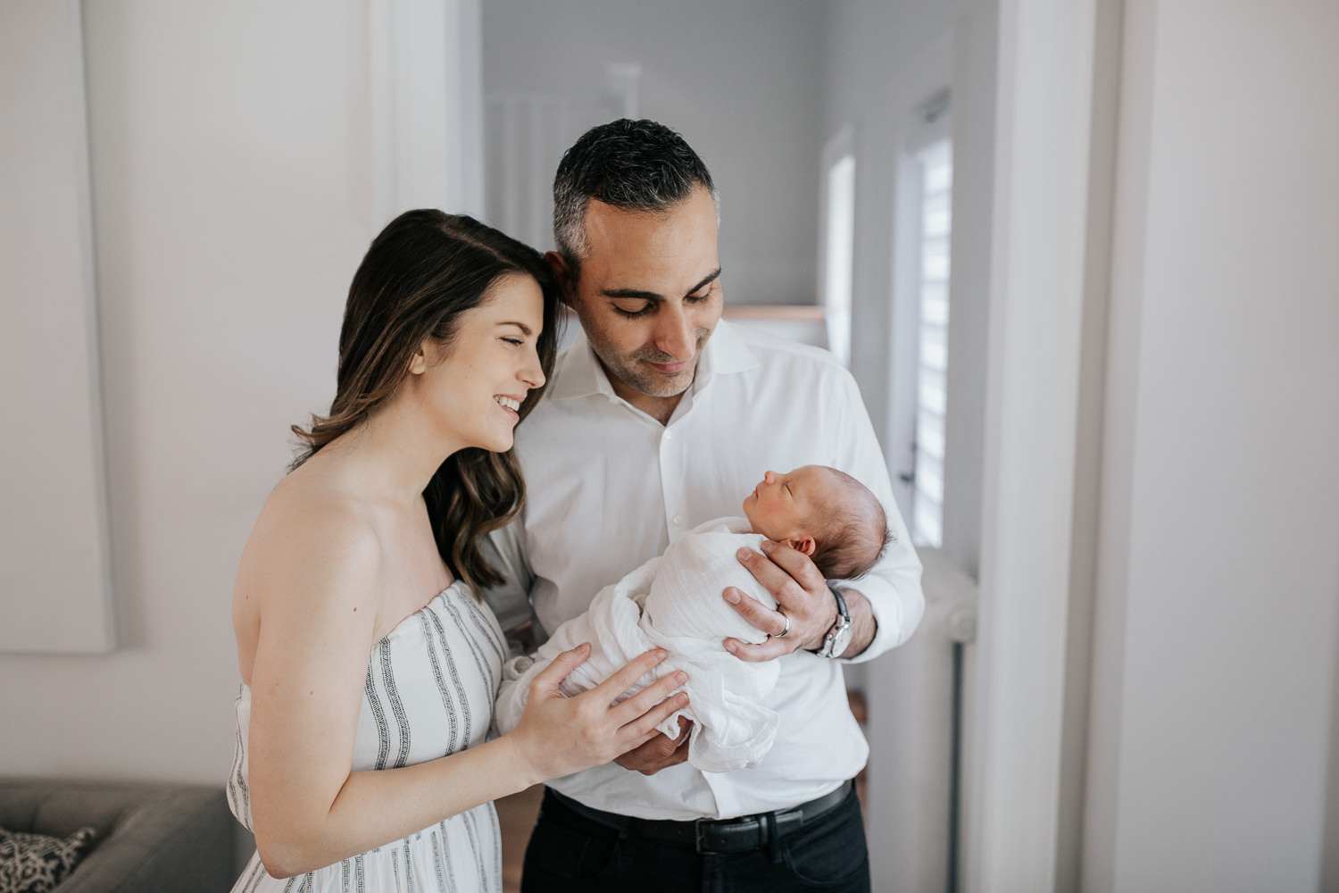 new parents standing in living room, father holding sleeping, swaddled 2 week old baby boy in his arms, mother standing next to husband, smiling down at son - Stouffville In-Home Photography