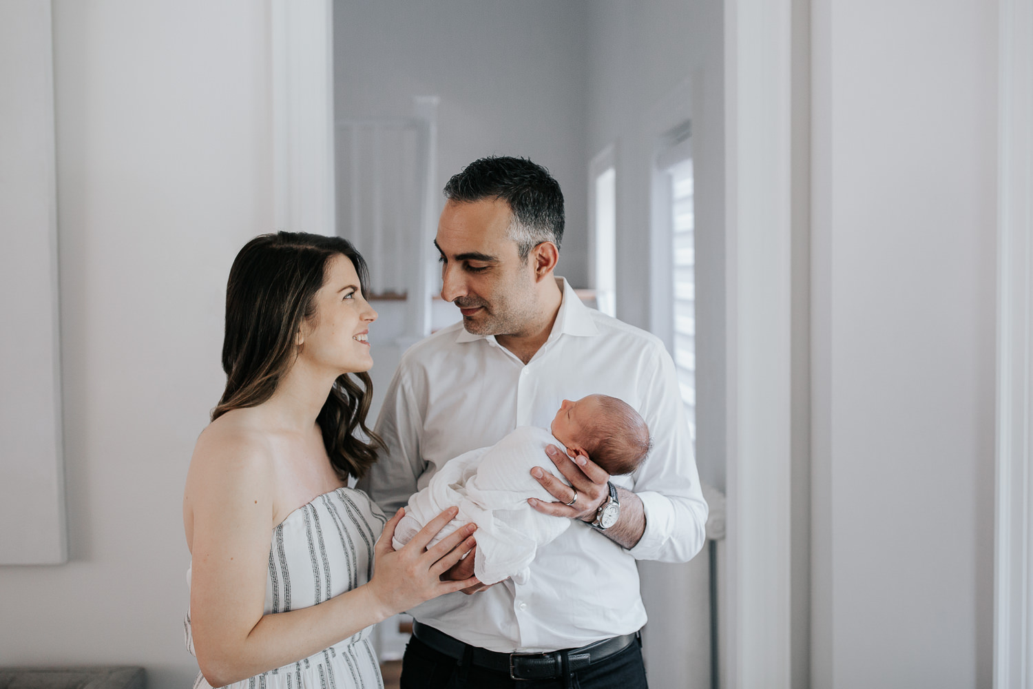 new parents standing in living room smiling at each other, father holding sleeping, swaddled 2 week old baby boy in his arms, mother standing next to husband - York Region In-Home Photography