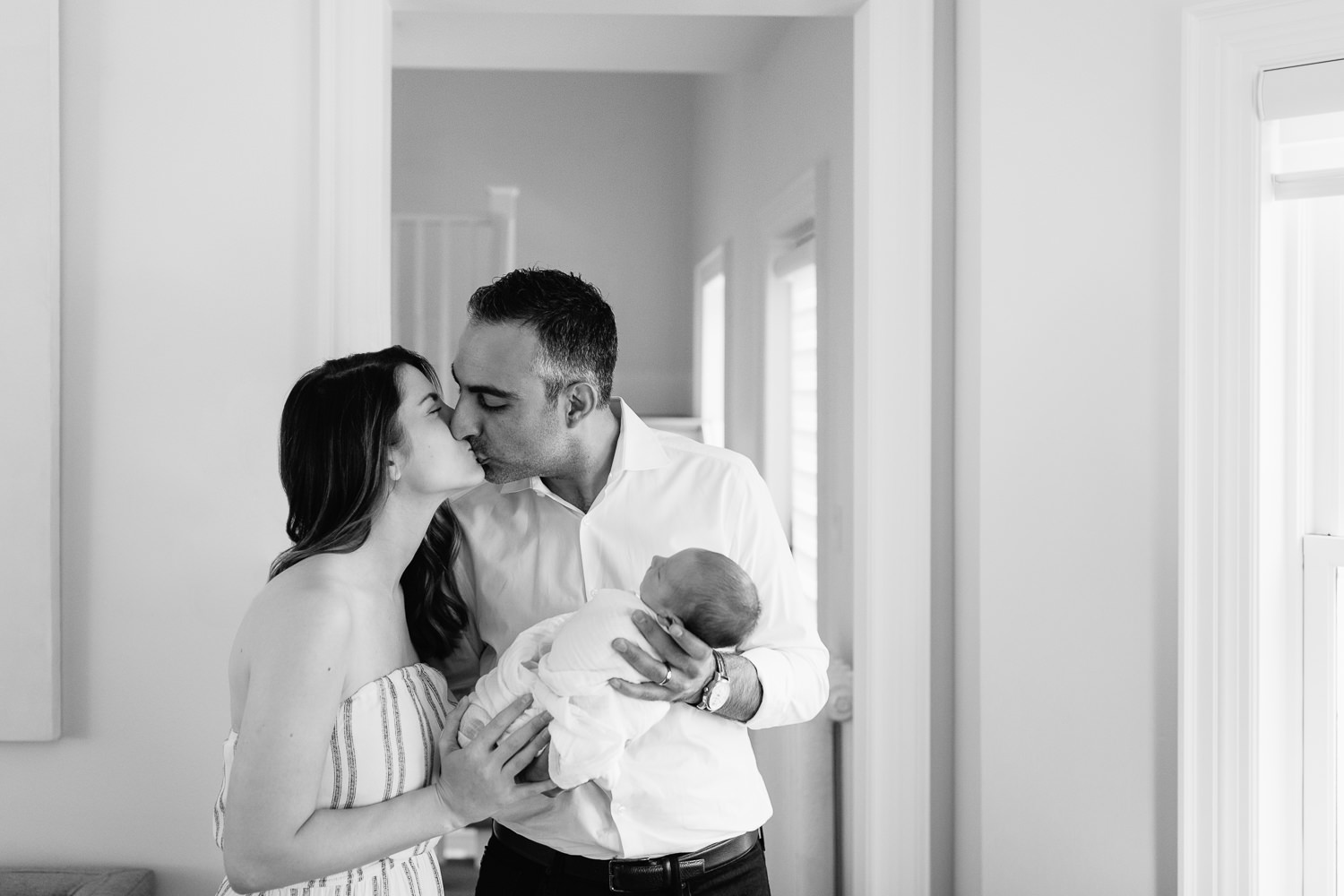 new parents standing in living room, father holding sleeping, swaddled 2 week old baby boy in his arms, mother standing next to them, husband and wife kissing - Markham In-Home Photography