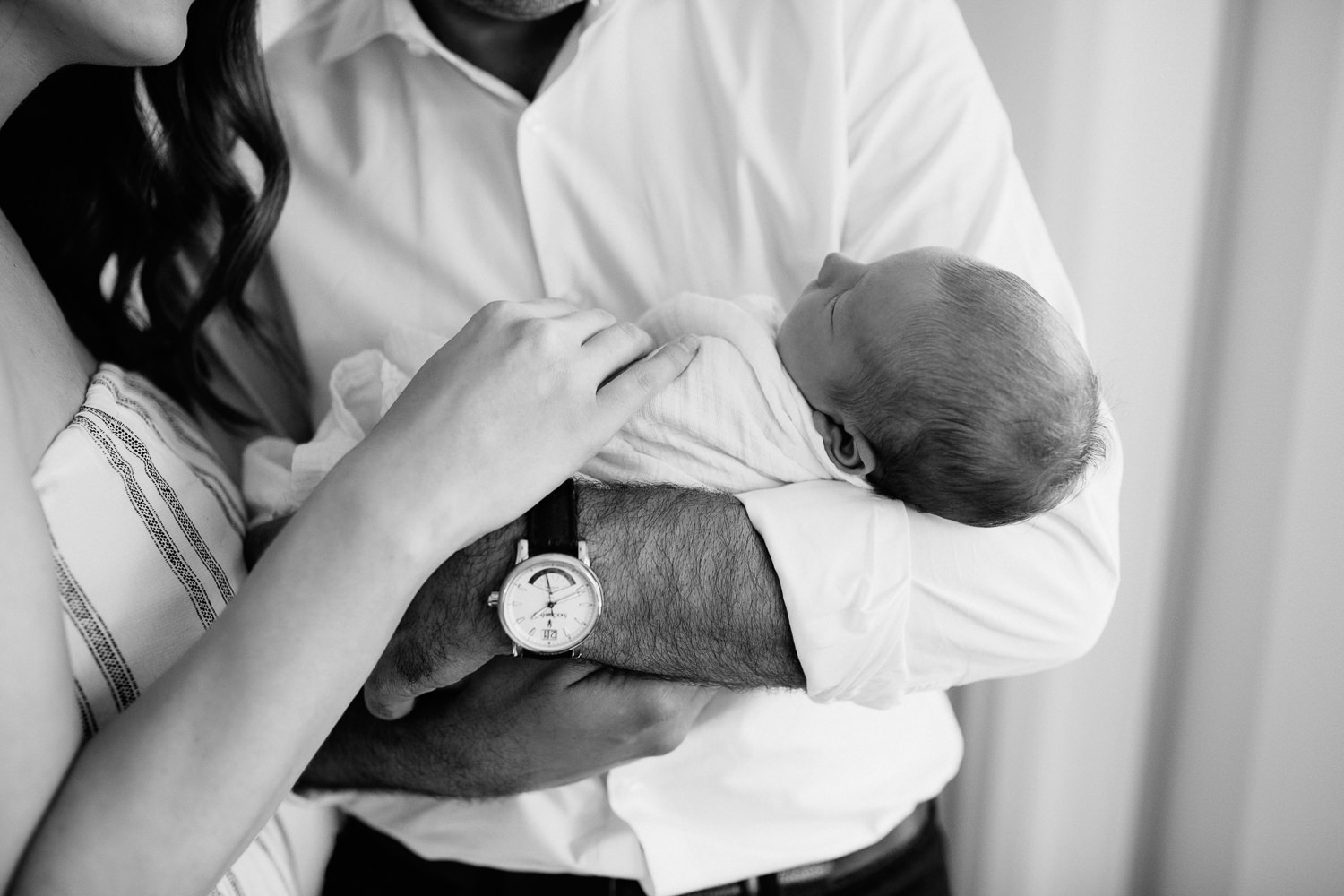 new parents standing in living room, father holding sleeping, swaddled 2 week old baby boy in his arms, mother standing next to husband, hand on son - GTA In-Home Photography