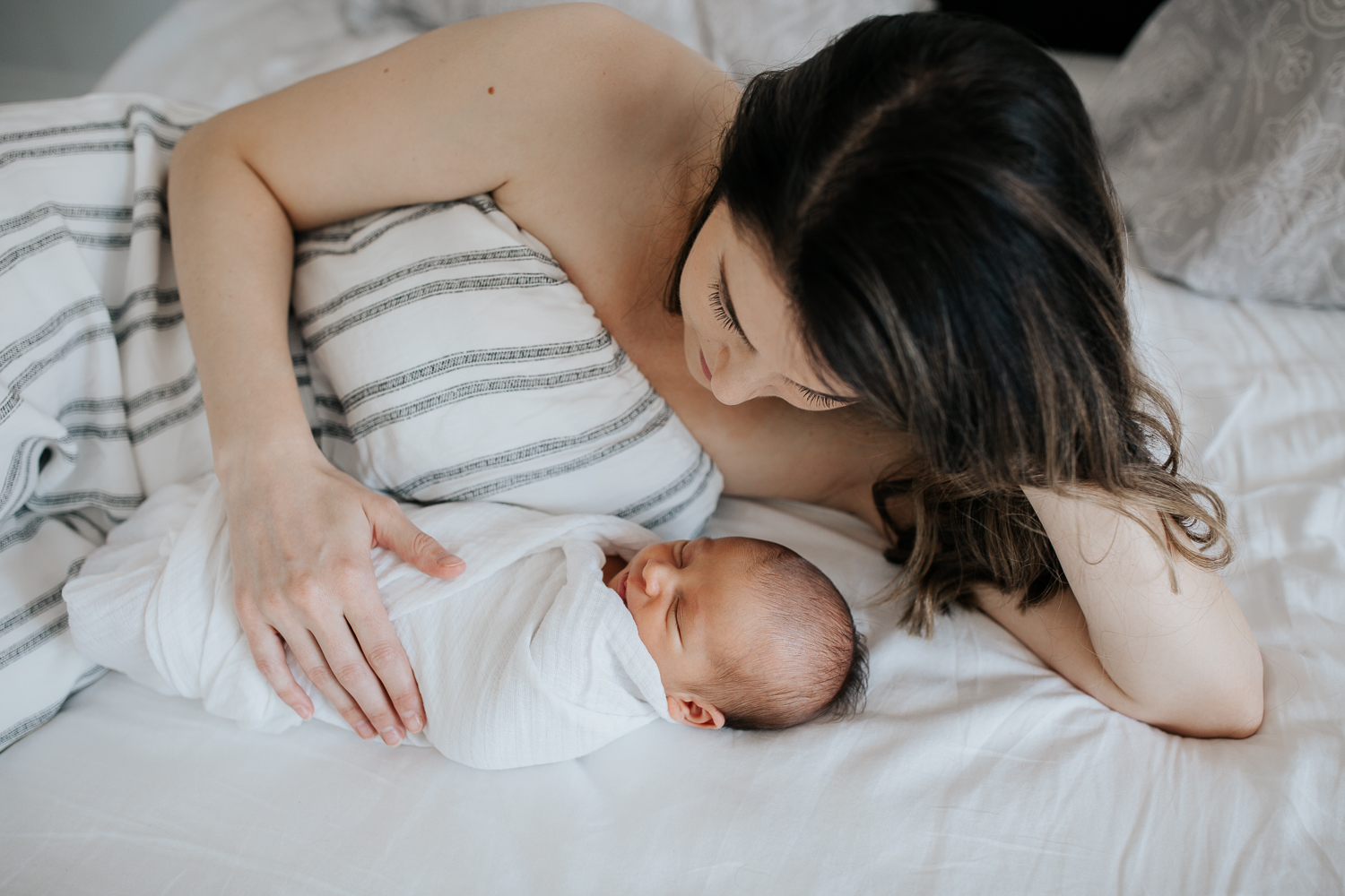 new mother with long dark hair lying on bed next to sleeping 2 week old baby boy wrapped in white swaddle - Stouffville Lifestyle Photos