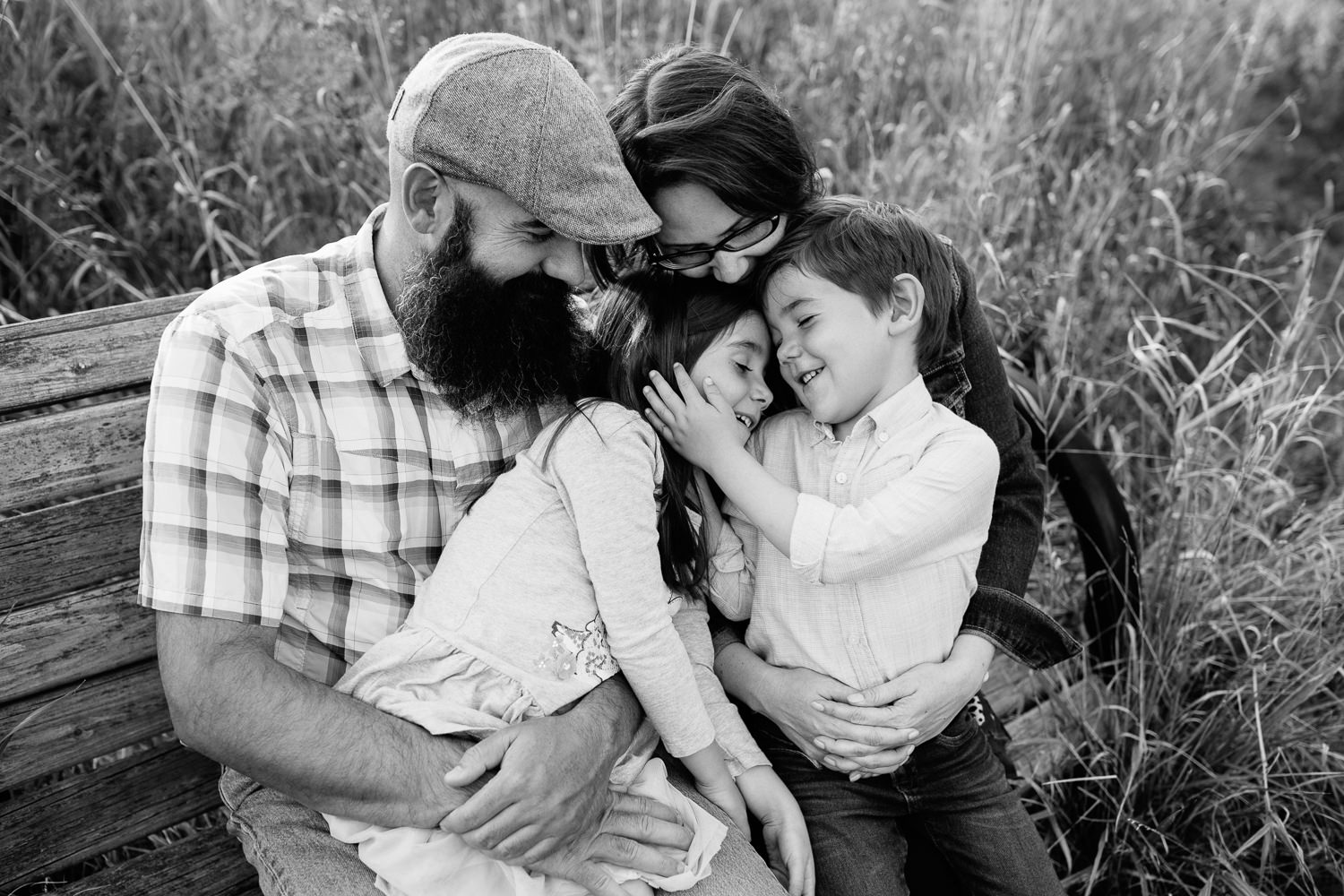 family of 4 sitting on park bench surrounded by tall grasses, 5 year old girl sitting on dad's lap, boy in mom's, snuggling, brother's hand on sister's face - Newmarket Lifestyle Photography