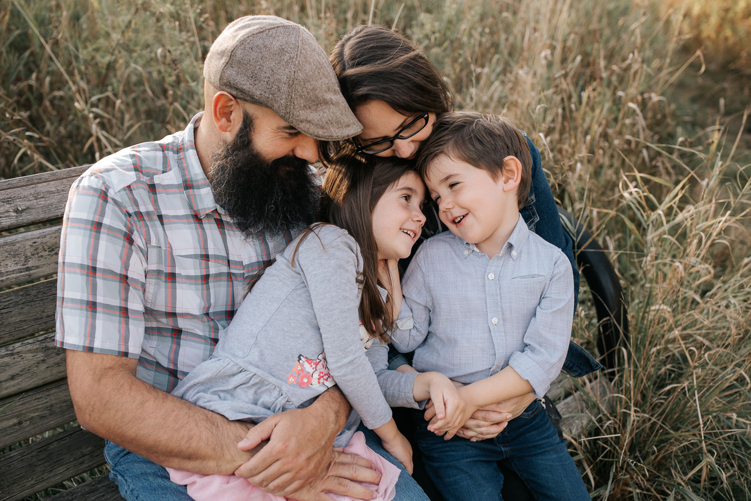 family of 4 sitting on park bench surrounded by tall grasses, 5 year old girl sitting on dad's lap, boy in mom's, snuggling, brother looking at sister - Stouffville Lifestyle Photography