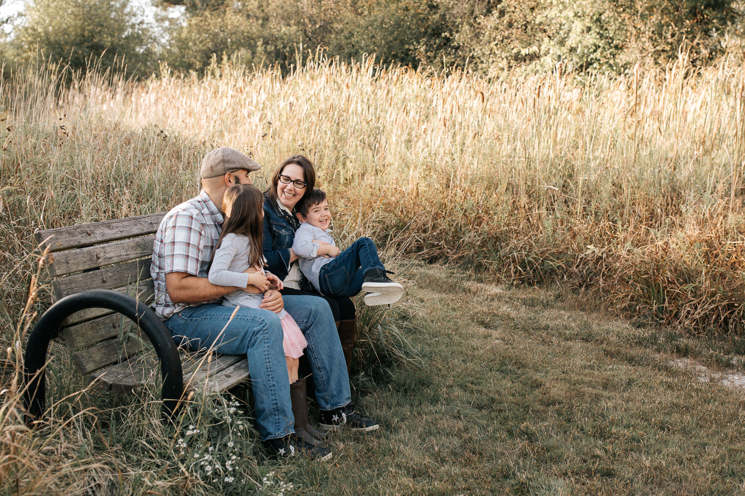 family of 4 sitting on park bench surrounded by tall grasses, 5 year old girl sitting on dad's lap, 4 year old boy in mom's lap smiling - Barrie Lifestyle Photography