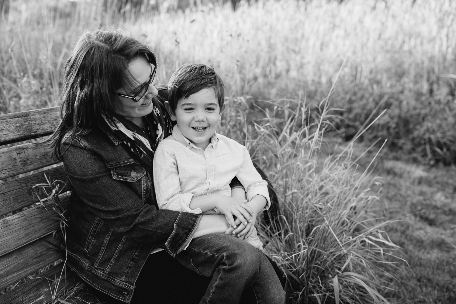 mother with dark brown hair sitting on park bench surrounded by tall grasses with 4 year old boy sitting in her lap, mom smiling at son as he smiles - Newmarket In-Home Photography