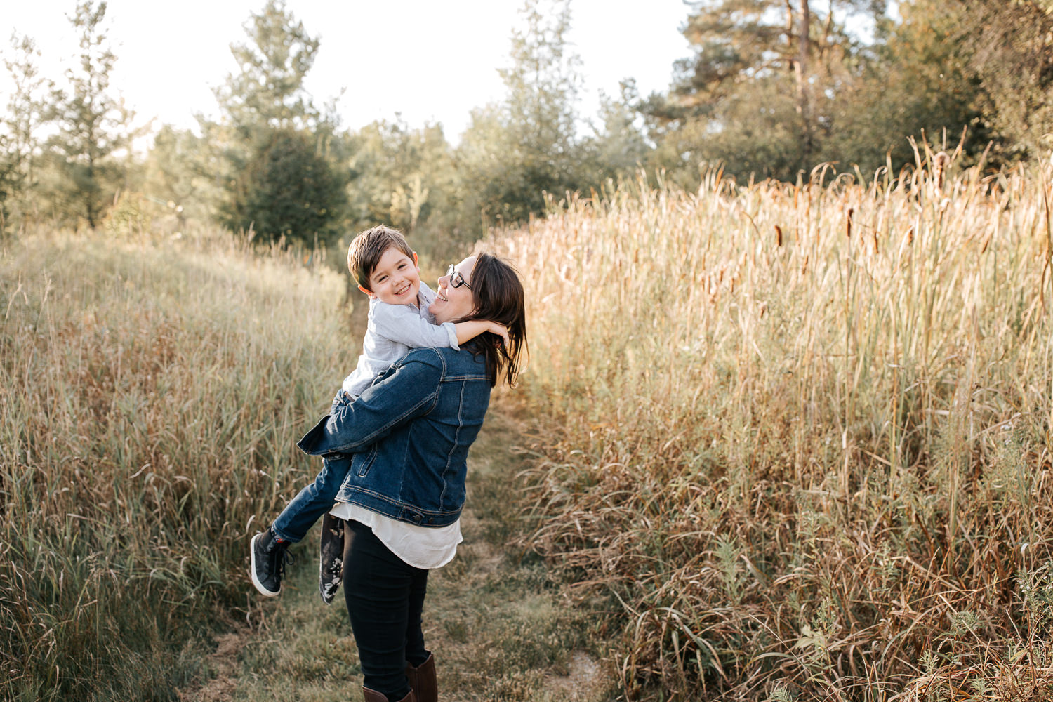 mother with dark brown hair standing in field surrounded by tall grasses with 4 year old boy in her arms, son smiling at camera - Stouffville In-Home Photography