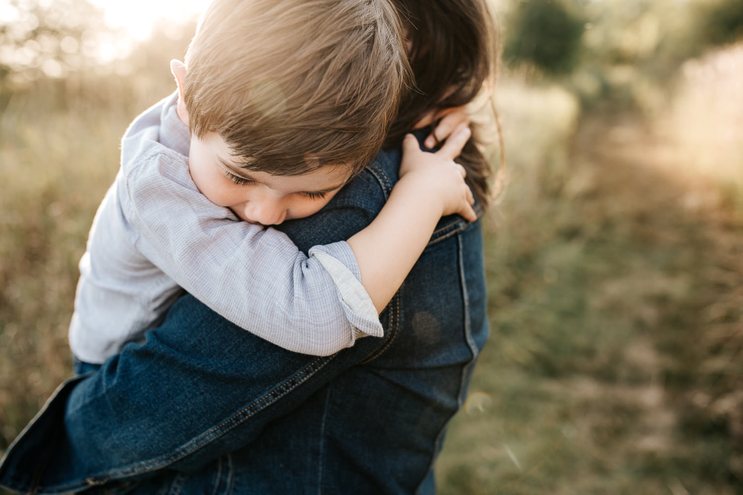 mother with dark brown hair standing in field surrounded by tall grasses, holding 4 year old boy, son with arm around mom's neck and head on her shoulder - Markham In-Home Photography