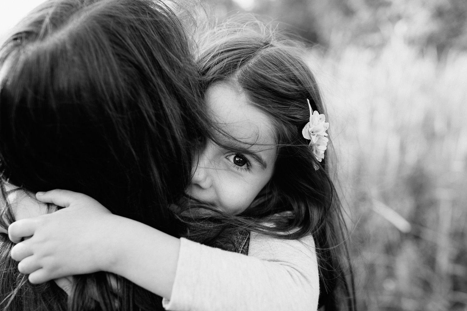 mother with dark brown hair standing in field surrounded by tall grasses, holding 5 year old girl, daughter with arms around mom's neck looking at camera over her shoulder - Barrie In-Home Photography