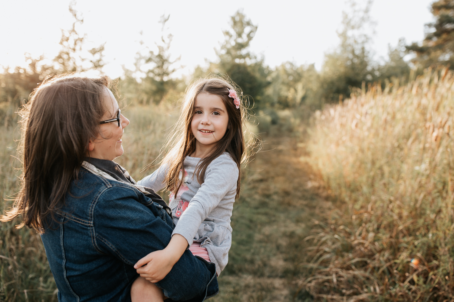 mother with dark brown hair standing in field surrounded by tall grasses with 5 year old girl in her arms, daughter looking at camera - GTA In-Home Photography