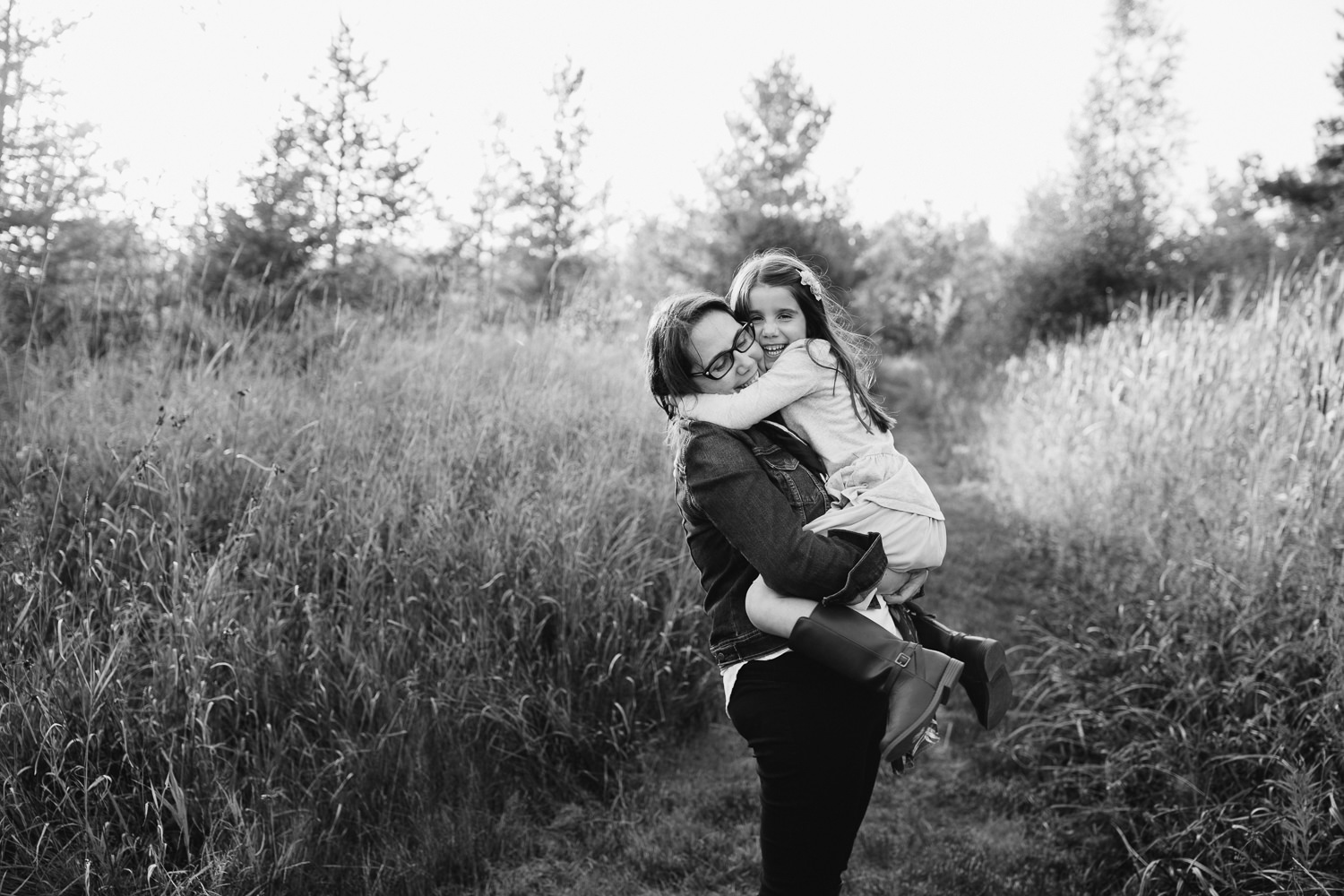 mother with dark brown hair standing in field surrounded by tall grasses with 5 year old girl in her arms, daughter hugging mom and looking at camera - York Region In-Home Photography