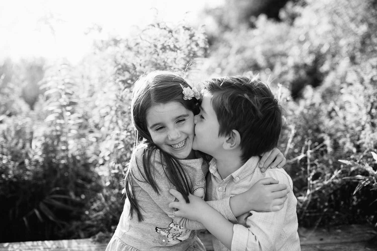 5 year old girl and 4 year old boy with dark hair sit on bench, sister hugging brother and smiling as he kisses her on cheek - Newmarket In-Home Photos