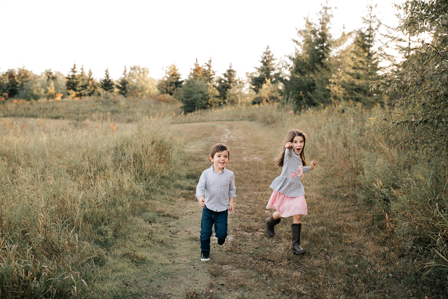 4 year old boy and 5 year old girl with dark hair running down path in grassy field smiling, sister's arms outstretched towards camera - Newmarket Lifestyle Photos