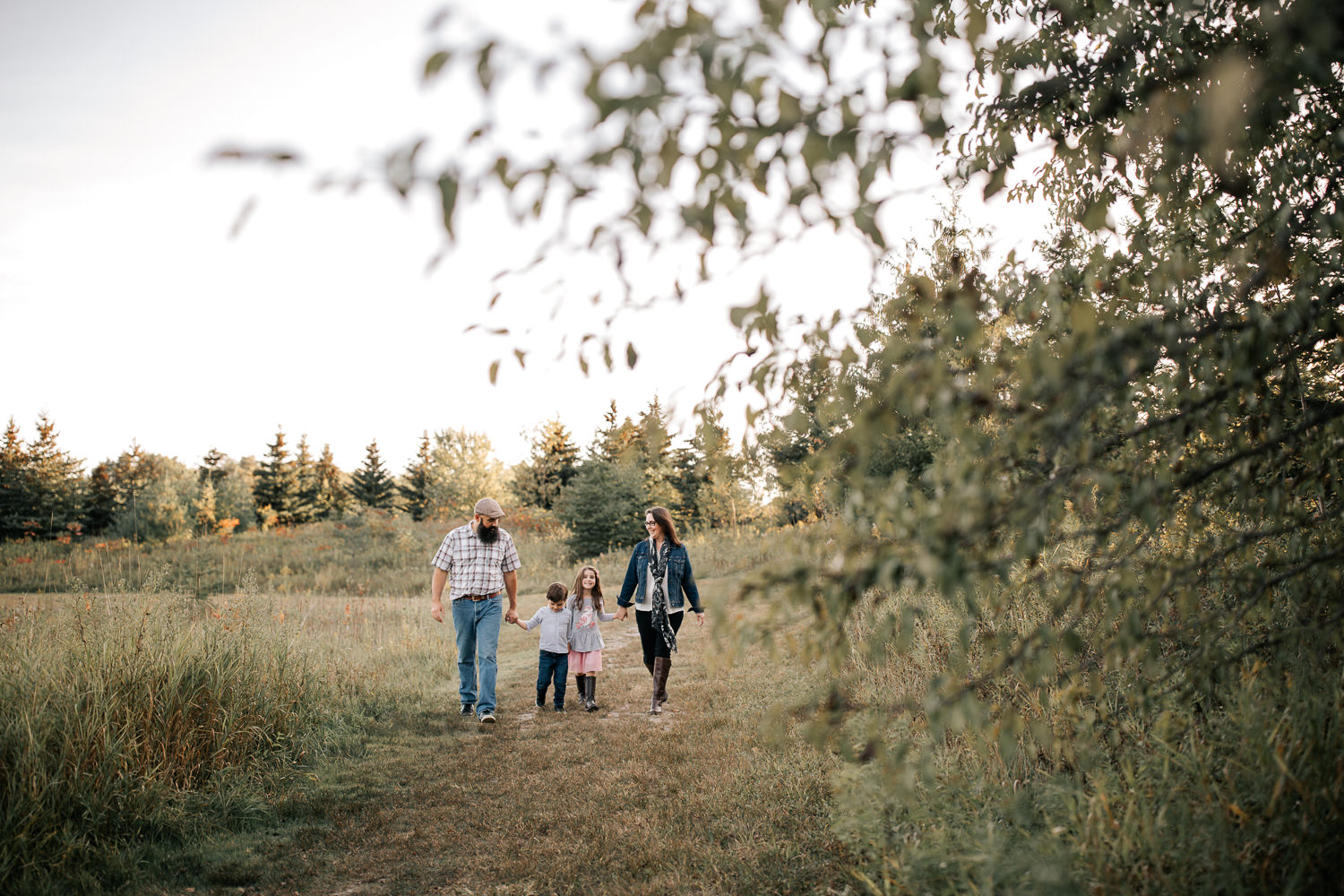 family of 4 holding hands, walking down path in grassy field, smiling at each other, trees in foreground - Barrie Lifestyle Photos