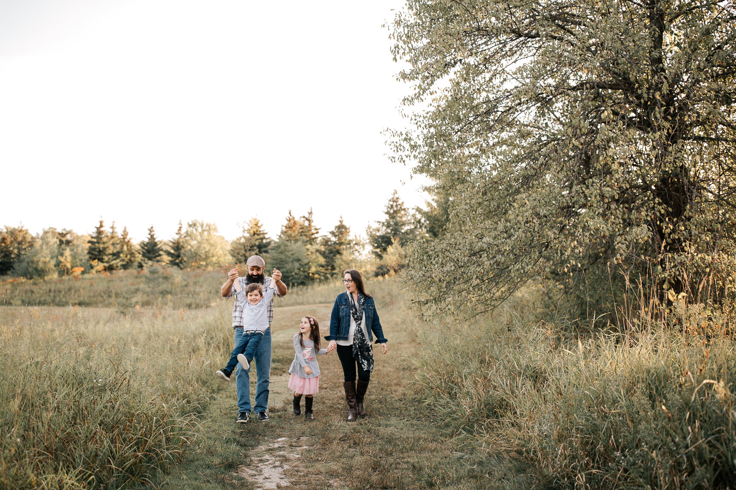 family of 4 holding hands, walking down path in grassy field, dad swinging son by both arms, mother and daughter laughing at them - York Region Lifestyle Photos