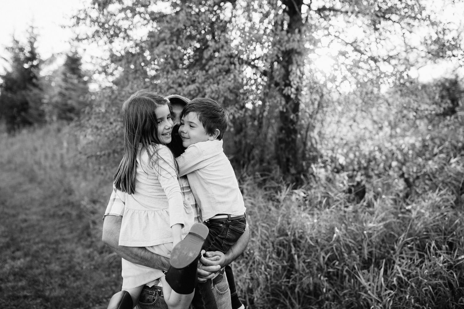 father standing in field holding 5 year old girl and 4 year old boy in his arms as daughter and son hug him - Markham Lifestyle Photography