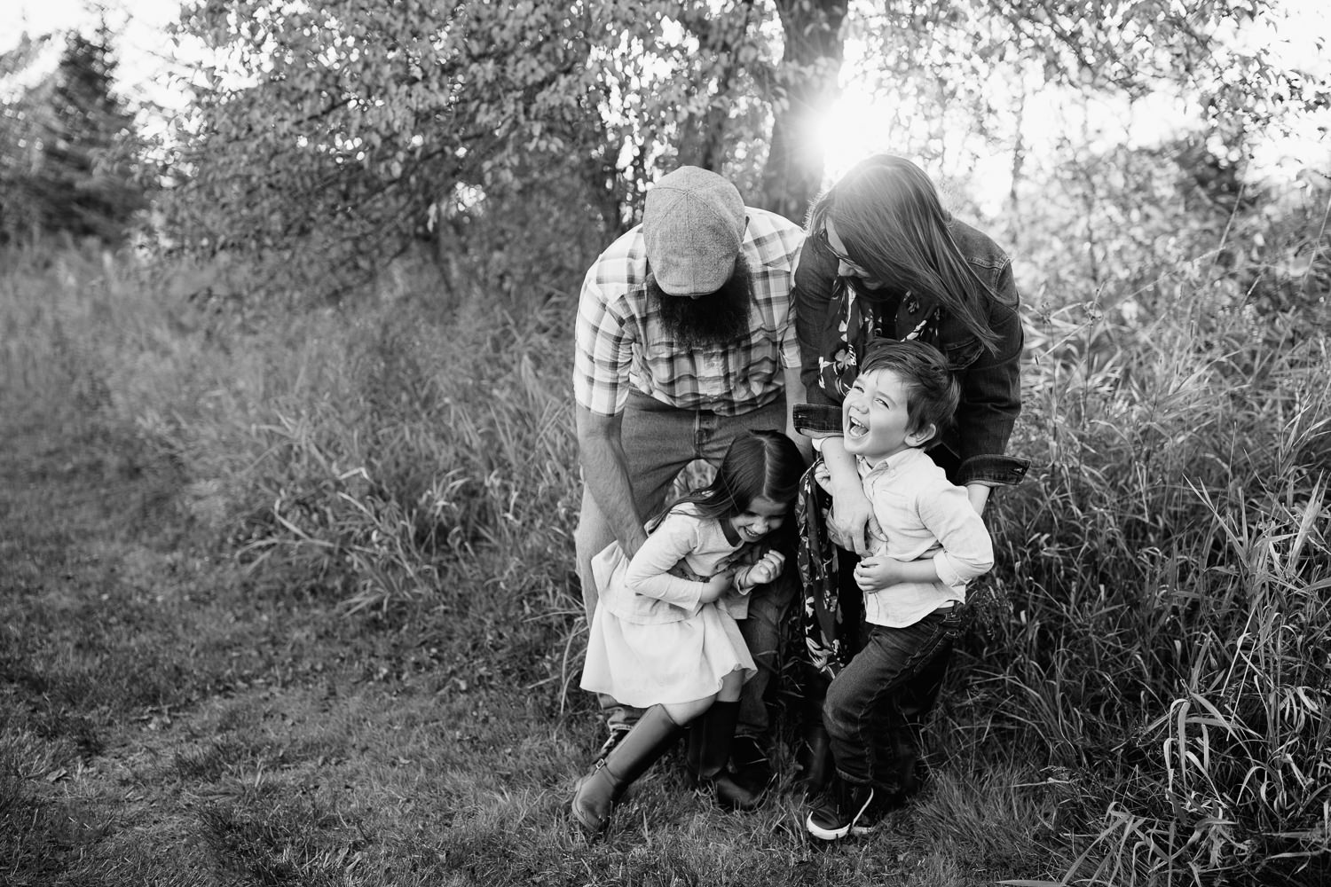 family of 4 standing together in grass field in front of tree, mom and dad leaning over, father tickling 5 year old daughter and mother tickling son, kids laughing - Newmarket In-Home Photography