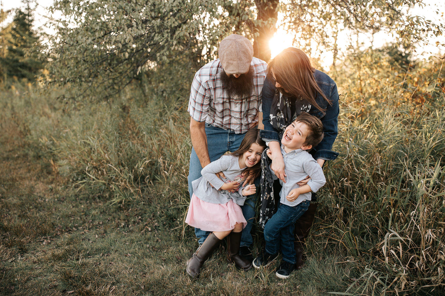 family of 4 standing together in grass field in front of tree, mom and dad leaning over, father tickling 5 year old daughter and mother tickling son, kids laughing - York Region Lifestyle Photography