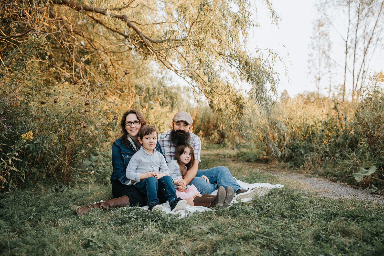 family of four sitting on blanket under willow tree, 4 year old boy sitting in mom's lap and 5 year old girl sitting in father's lap, everyone looking at camera - York Region In-Home Photography