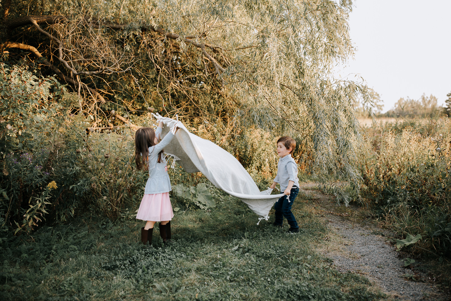5 year old girl and 4 year old boy standing in front of tree spreading white picnic blanket on the grass - Barrie In-Home Photography