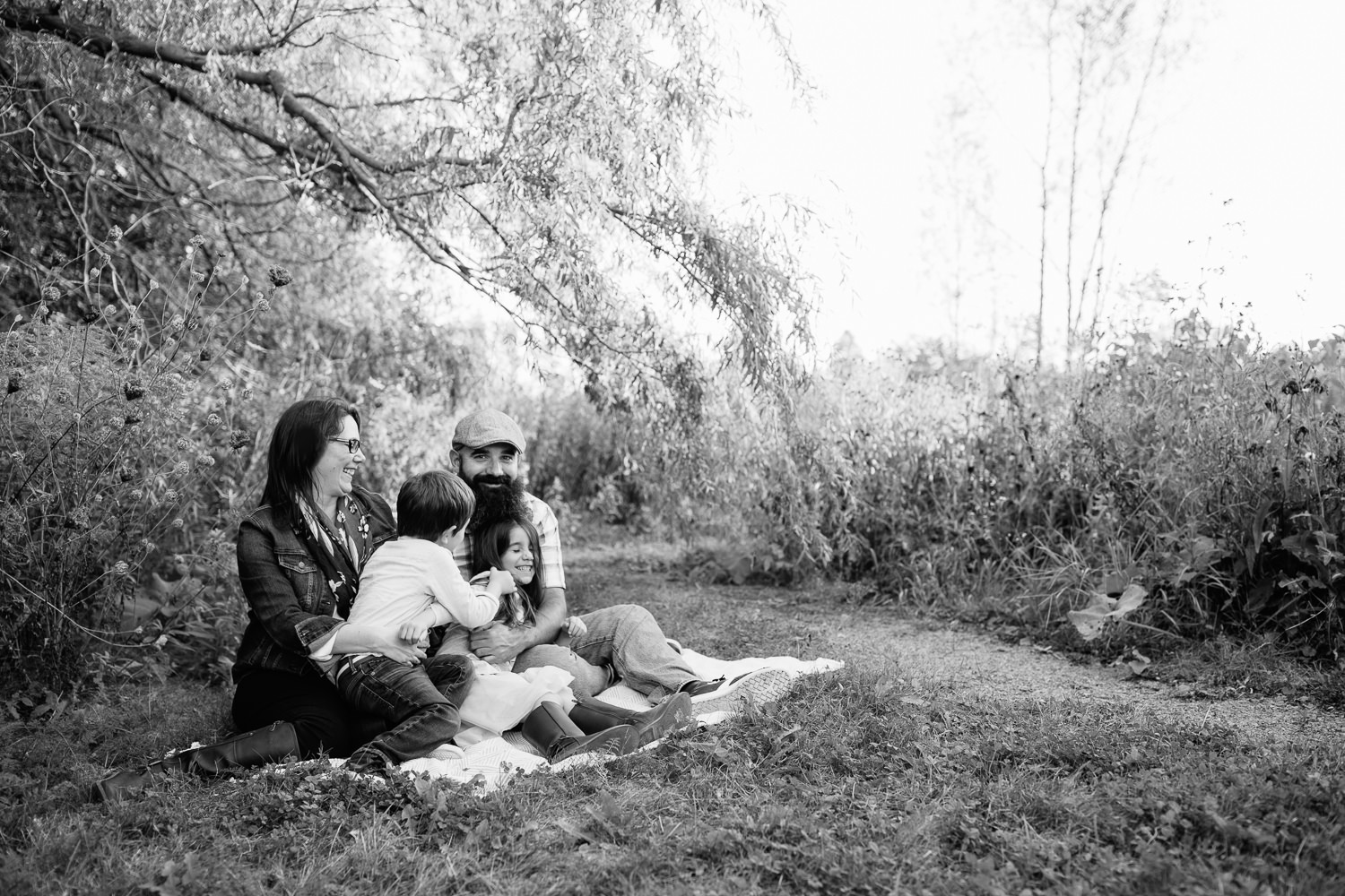 family of four sitting on blanket under willow tree, 4 year old boy sitting in mom's lap and leaning over to tickle big sister in dad's lap as she laughs -  GTA In-Home Photography