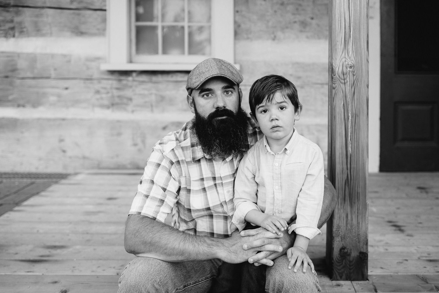 father with long dark beard sitting on wood porch step with 4 year old boy in his lap, dad's arms wrapped around son, both looking seriously at camera -Barrie In-Home Photos