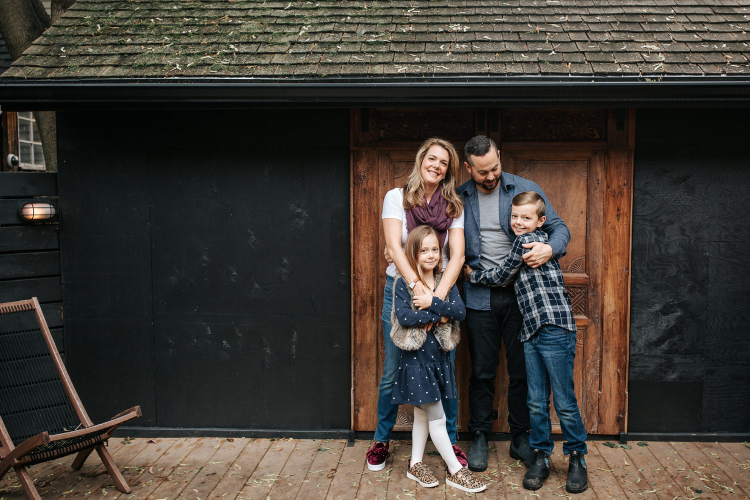 family of 4 standing on front of dark home with old doors, mom's arms around 7 year old girl's shoulders, both smiling at camera, 9 year old boy hugging dad - Newmarket Lifestyle Photography