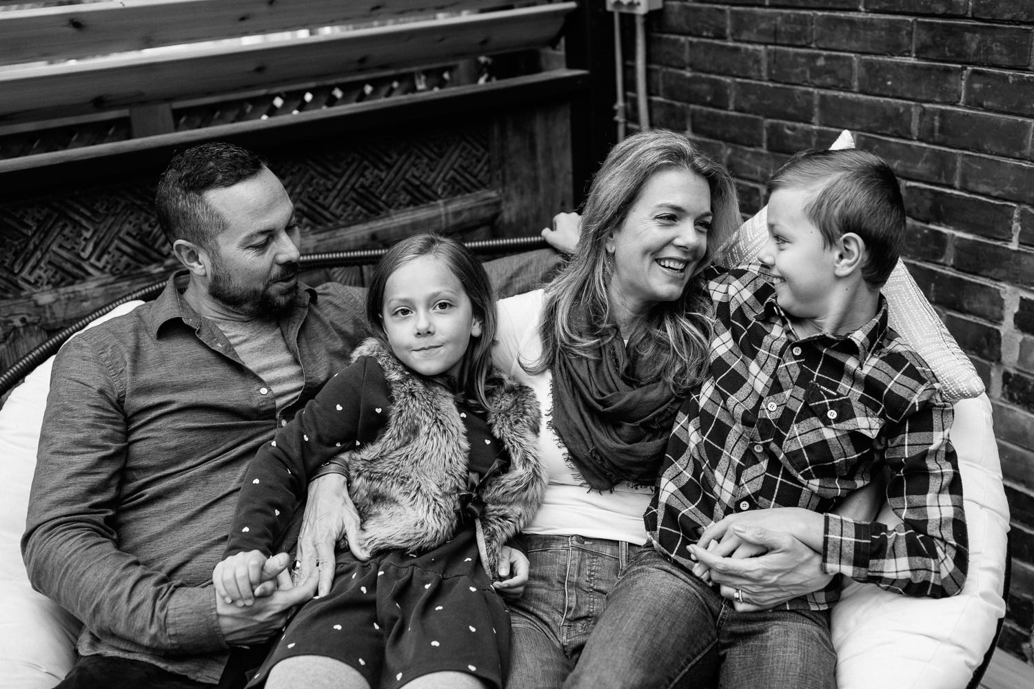 family of 4 sitting and snuggling on outdoor couch, 7 year old girl sitting in dad's lap holding his hand, 9 year old boy sitting in mom's lap smiling at her - GTA Lifestyle Photography