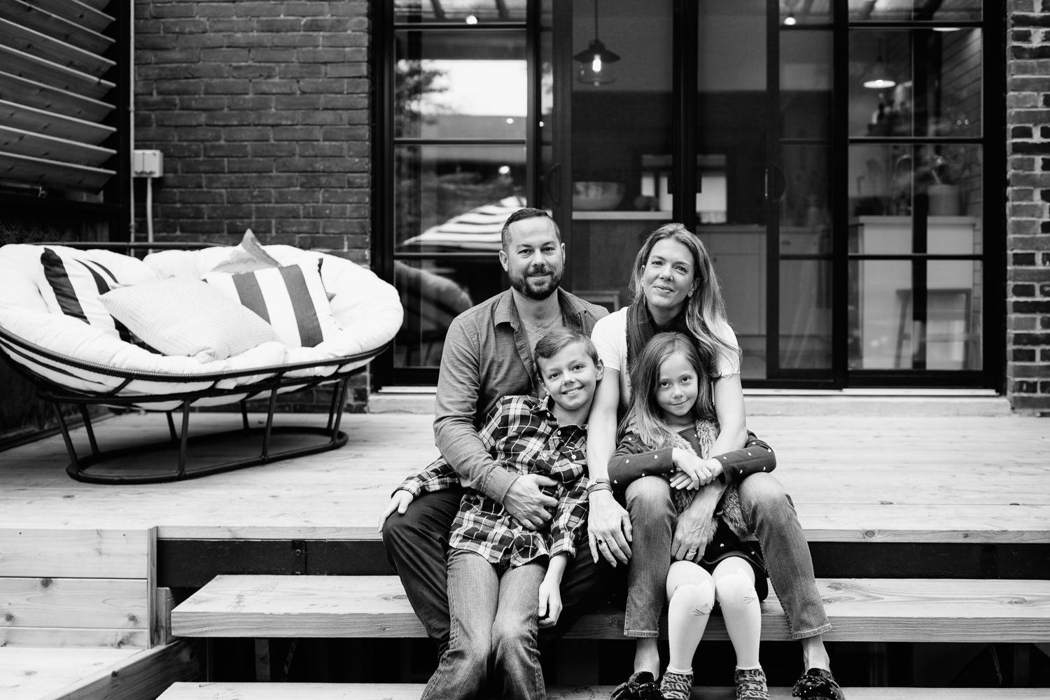 family of 4 sitting on backyard porch steps, 7 and 9 year old girl and boy sitting between parent's legs, everyone smiling at camera - Newmarket In-Home Photography