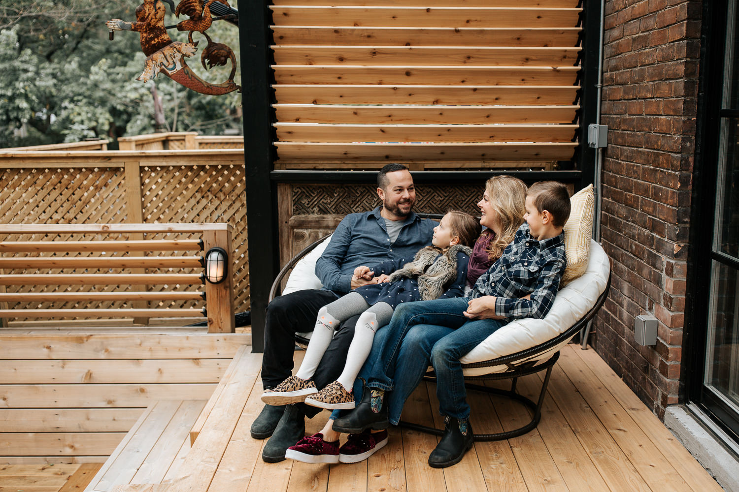 family of 4 sitting and snuggling on outdoor couch, 7 year old girl sitting in dad's lap holding his hand, 9 year old boy sitting in mom's, laughing and smiling - York Region Lifestyle Photography