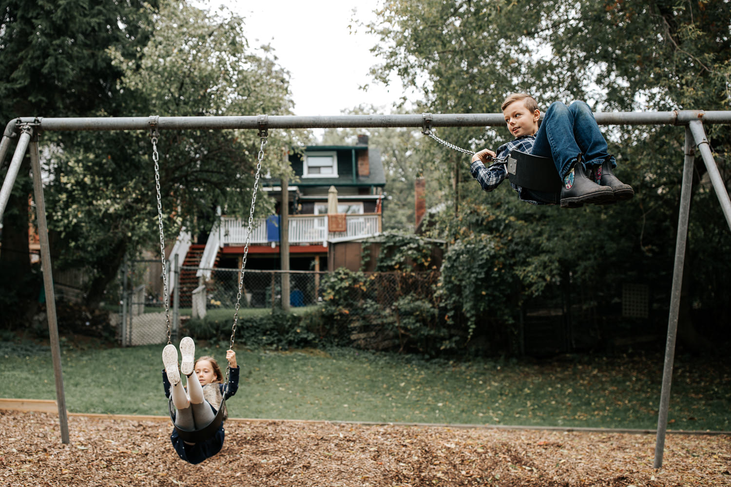 9 year old boy and 7 year old girl at outdoor park in the city swinging on swing set, brother swinging high in air and looking to the side - GTA In-Home Photography