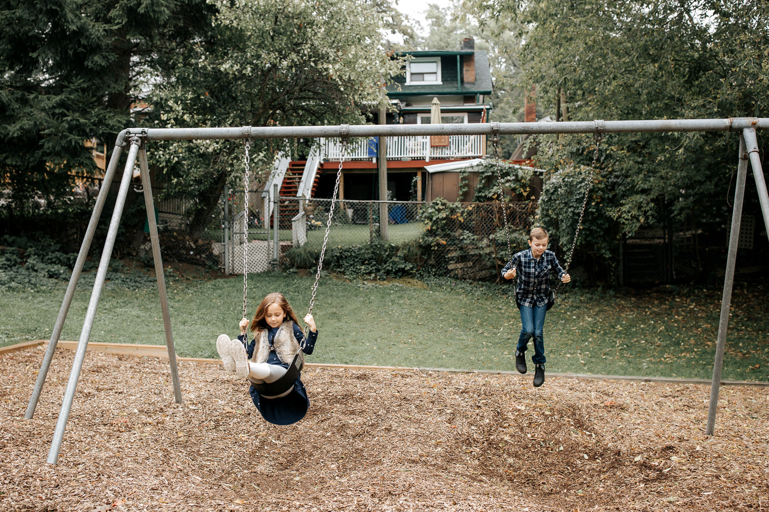 9 year old boy and 7 year old girl at outdoor park in the city swinging on swing set, focused looks on their faces - Barrie In-Home Photography