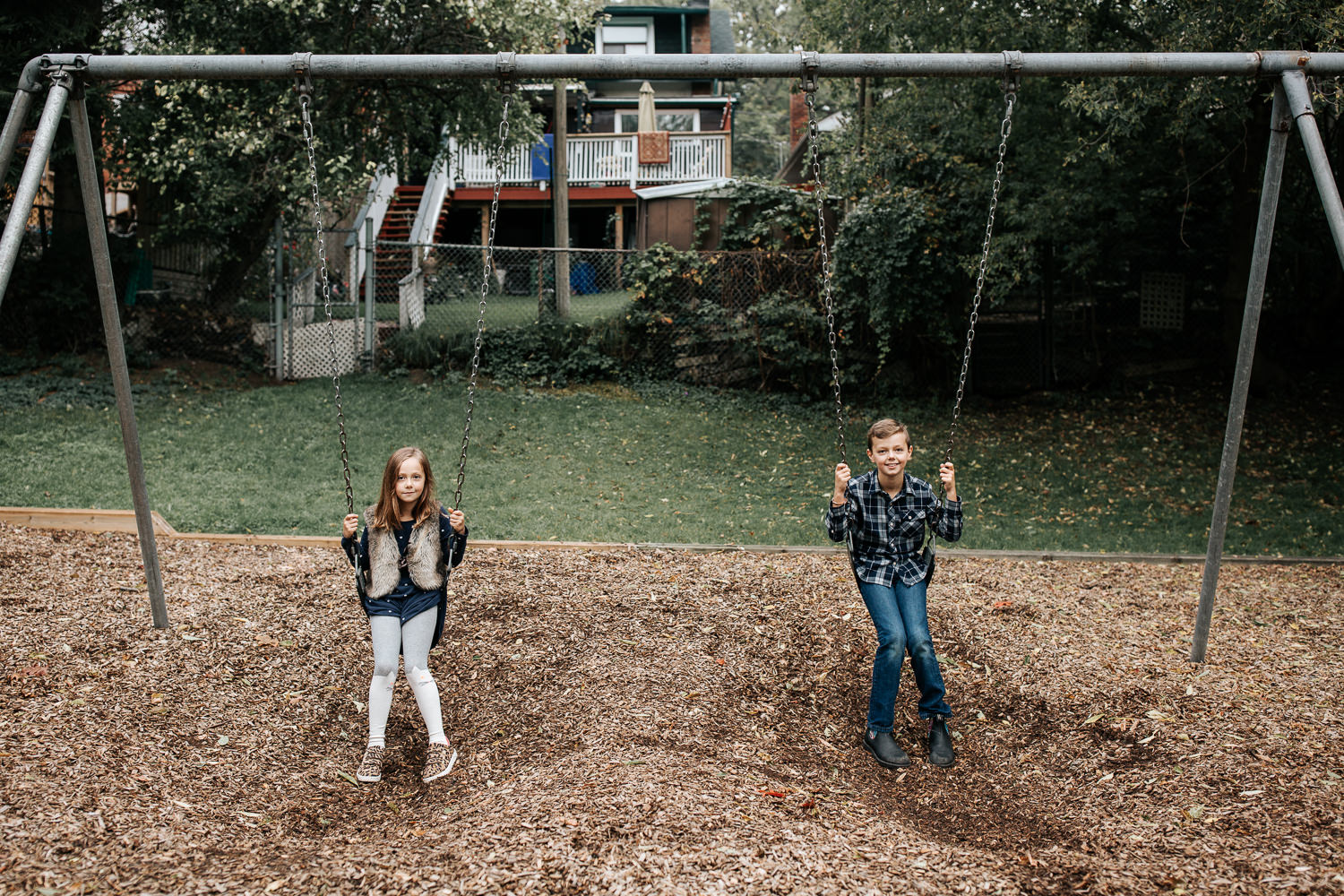 7 year old girl and 9 year old boy sitting still on swing set at city park, both looking  and smiling at the camera -  Stouffville In-Home Photos