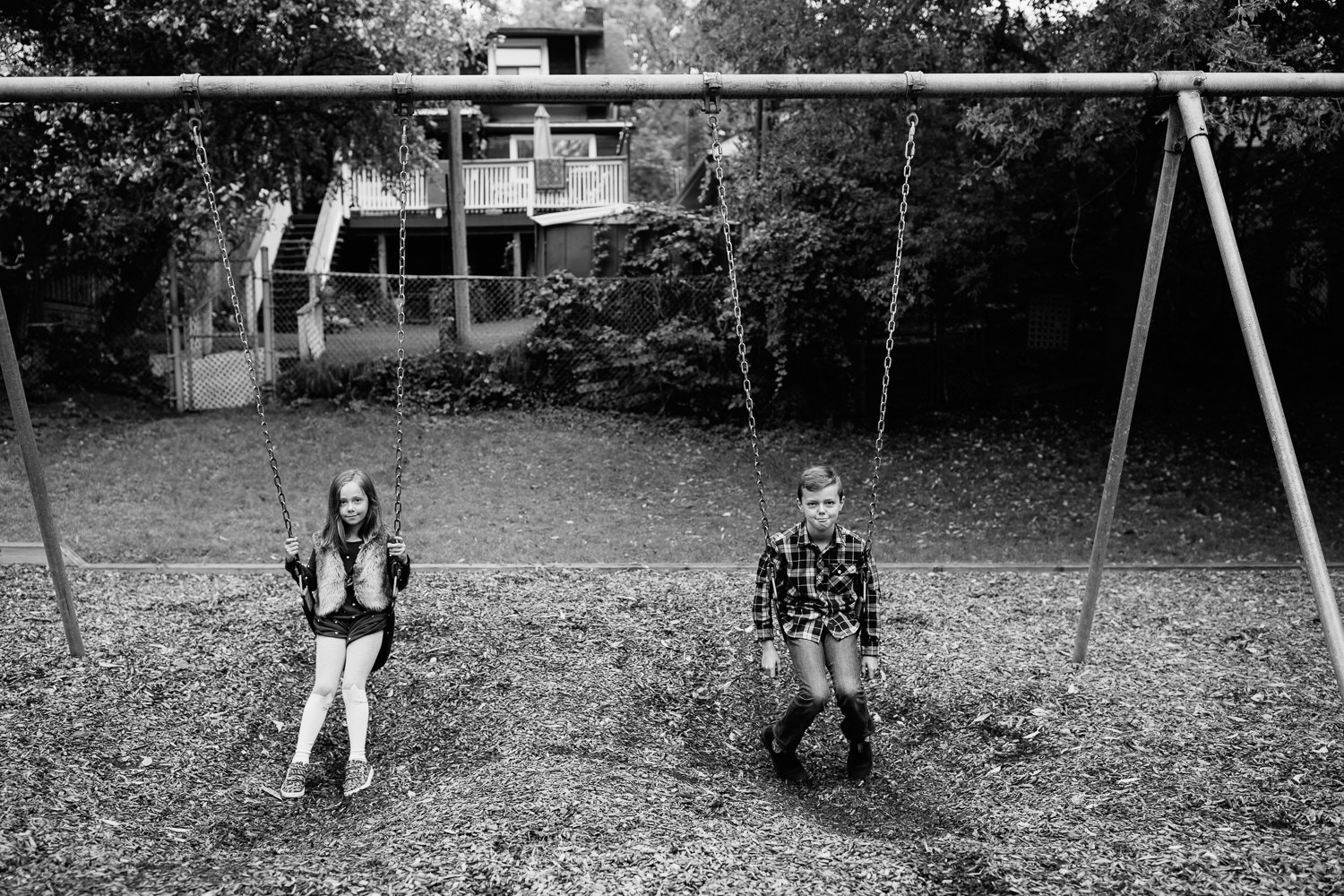 7 year old girl and 9 year old boy sitting still on swing set at city park, both looking seriously at the camera -  Markham In-Home Photos