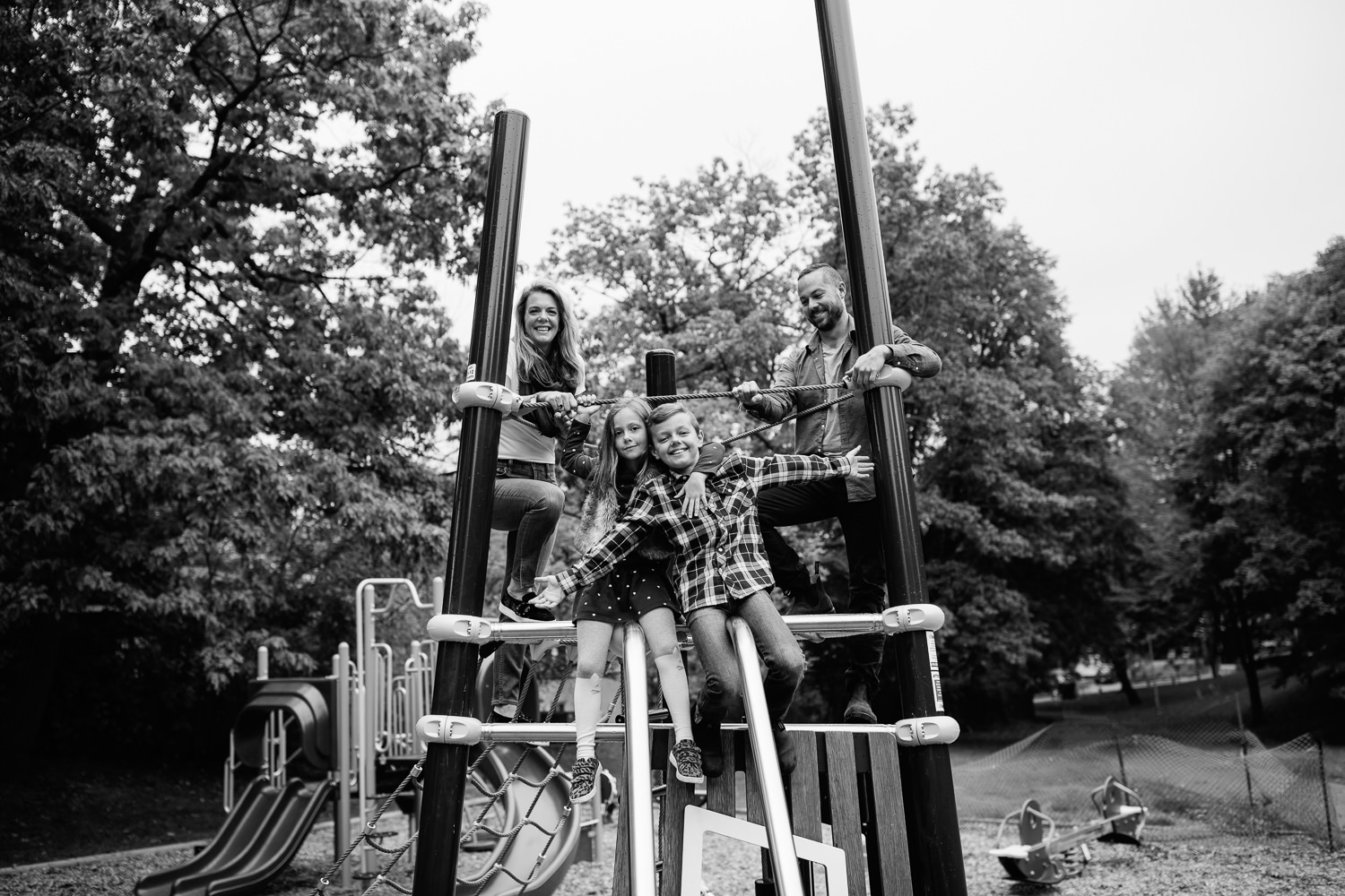 family of 4 standing on jungle gym at city park, 7 year old girl hugging brother, 9 year old boy with arms stretch out, mom and dad standing behind - Newmarket Lifestyle Photos