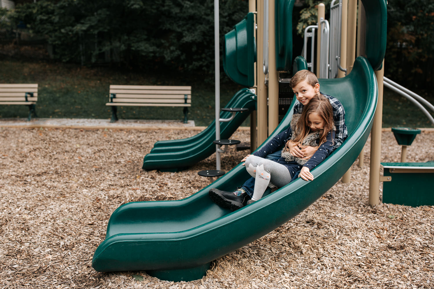 7 year old girl and 9 year old boy going down park slide, big brother sitting behind sister as they slide down - Barrie Lifestyle Photos