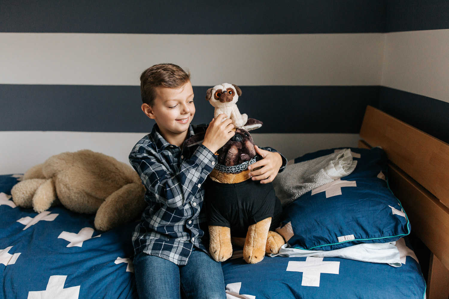 9 year old boy in plaid shirt with light brown hair sitting on bed in blue and white room holding toy pug stuffed animals, smiling at them - Markham In-Home Photography