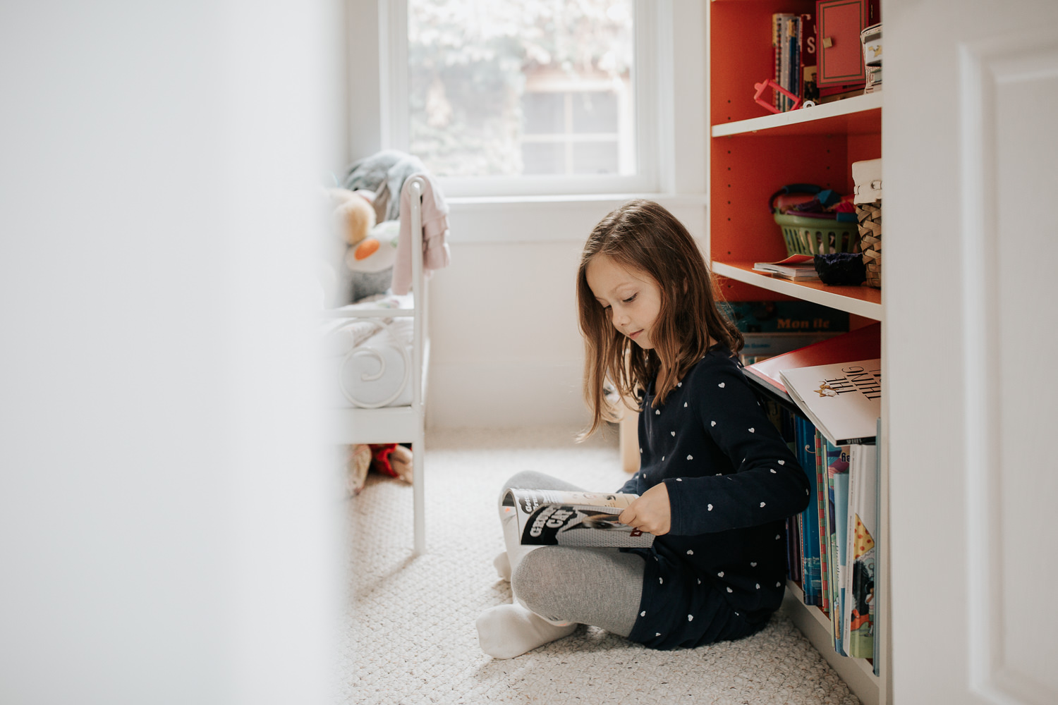 7 year old girl in polka dot dress with brown hair sitting on floor of bedroom leaning against orange and white bookcase and reading book - Stouffville In-Home Photos