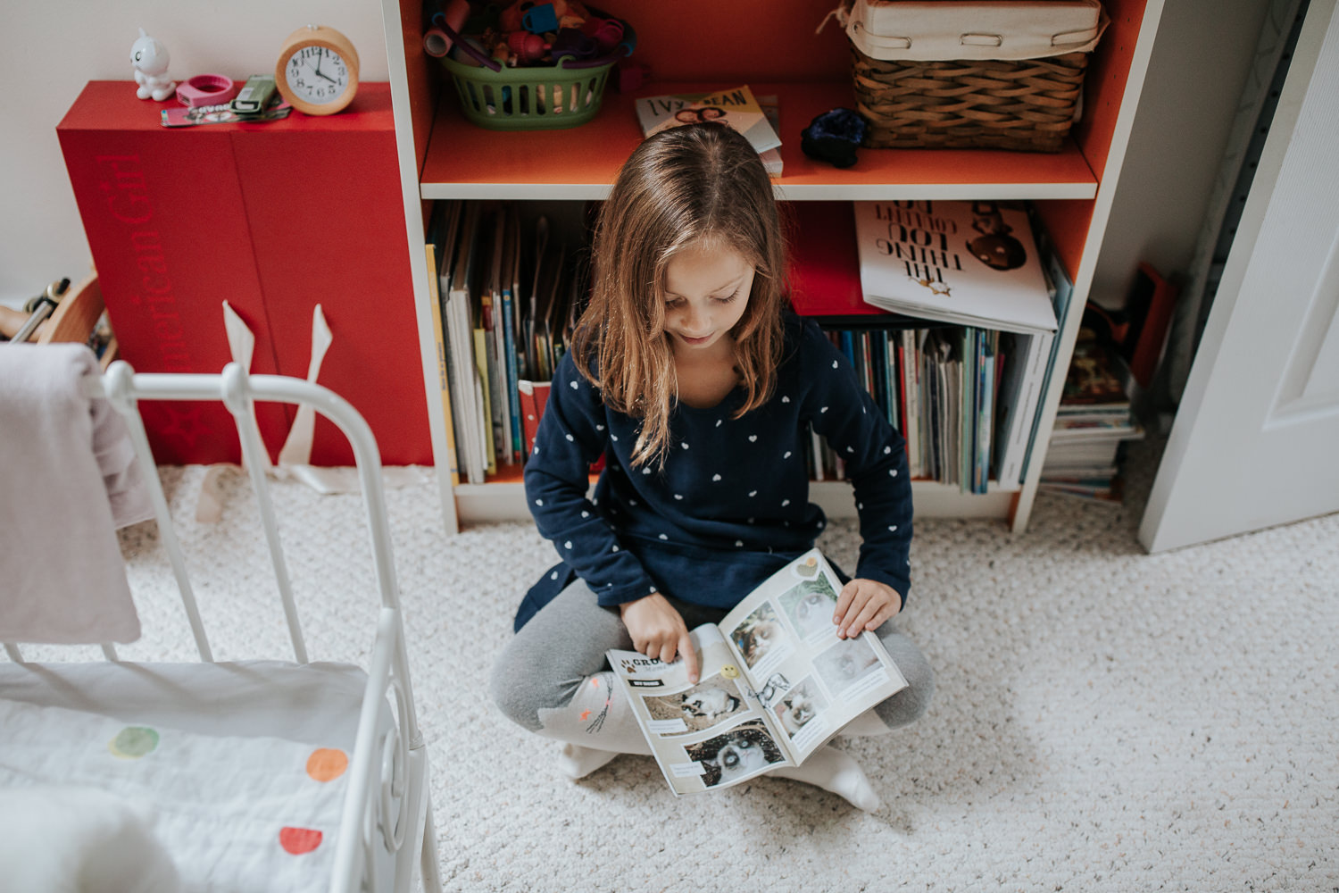 7 year old girl in polka dot dress with brown hair sitting on floor of bedroom leaning against orange and white bookcase and reading book - Markham In-Home Photos