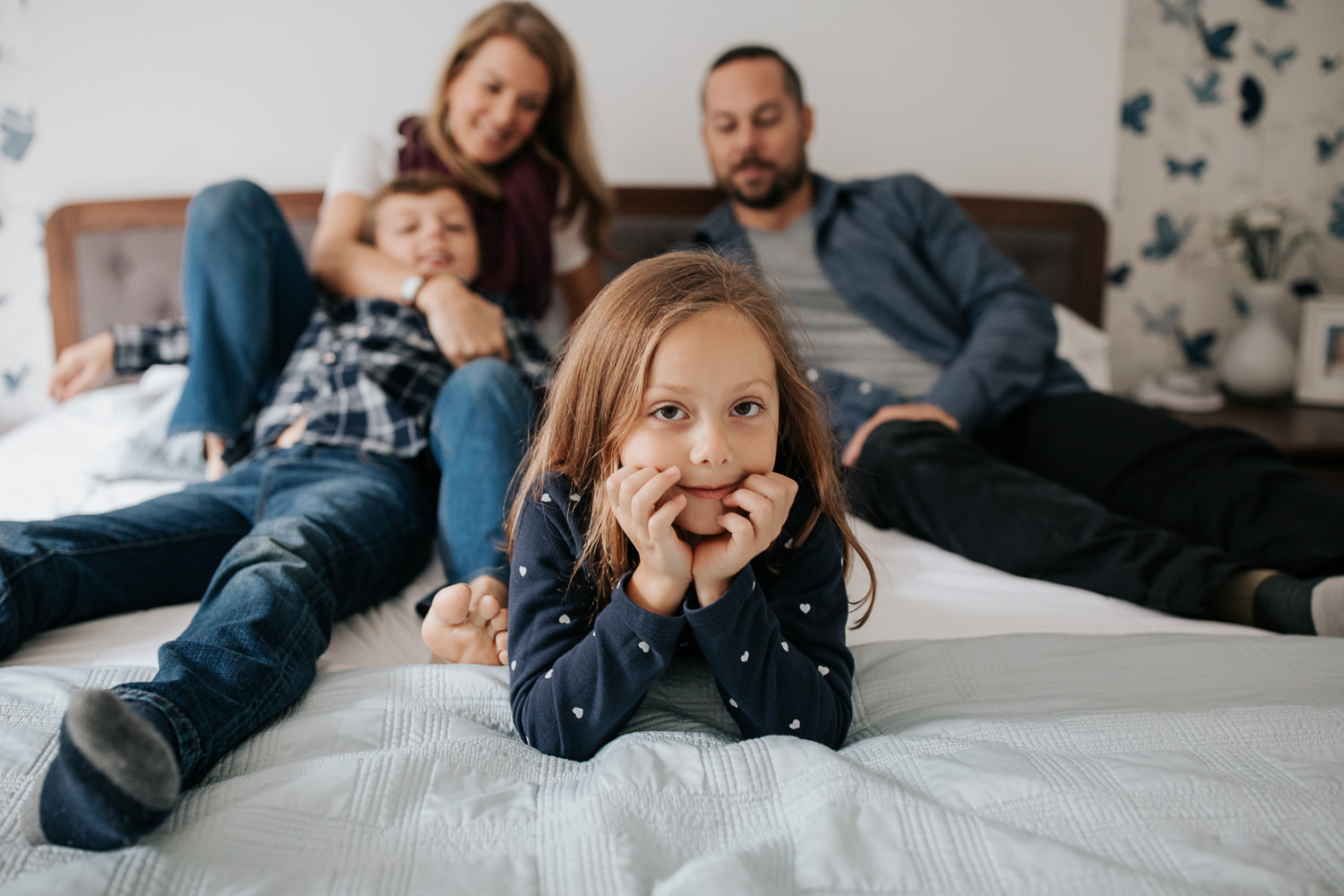 7 year old girl in polka dot dress lying on parents bed, head in hands looking at camera, and family sits on bed behind her - GTA In-Home Photos