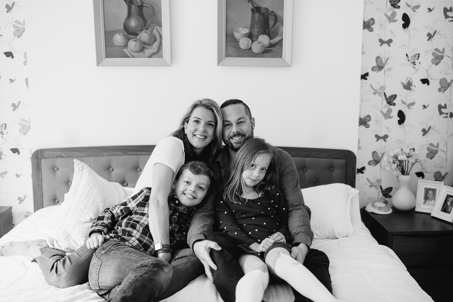 family of 4 sitting on master bed, 7 year old girl sitting in dad's lap, 9 year old boy in mom's everyone looking at camera, smiling - York Region In-Home Photos