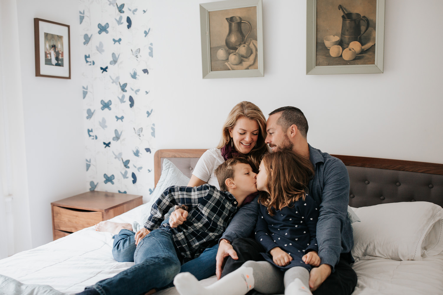family of 4 sitting on master bed, 7 year old girl sitting in dad's lap, 9 year old boy in mom's, brother giving little sister a kiss - Newmarket Lifestyle Photography