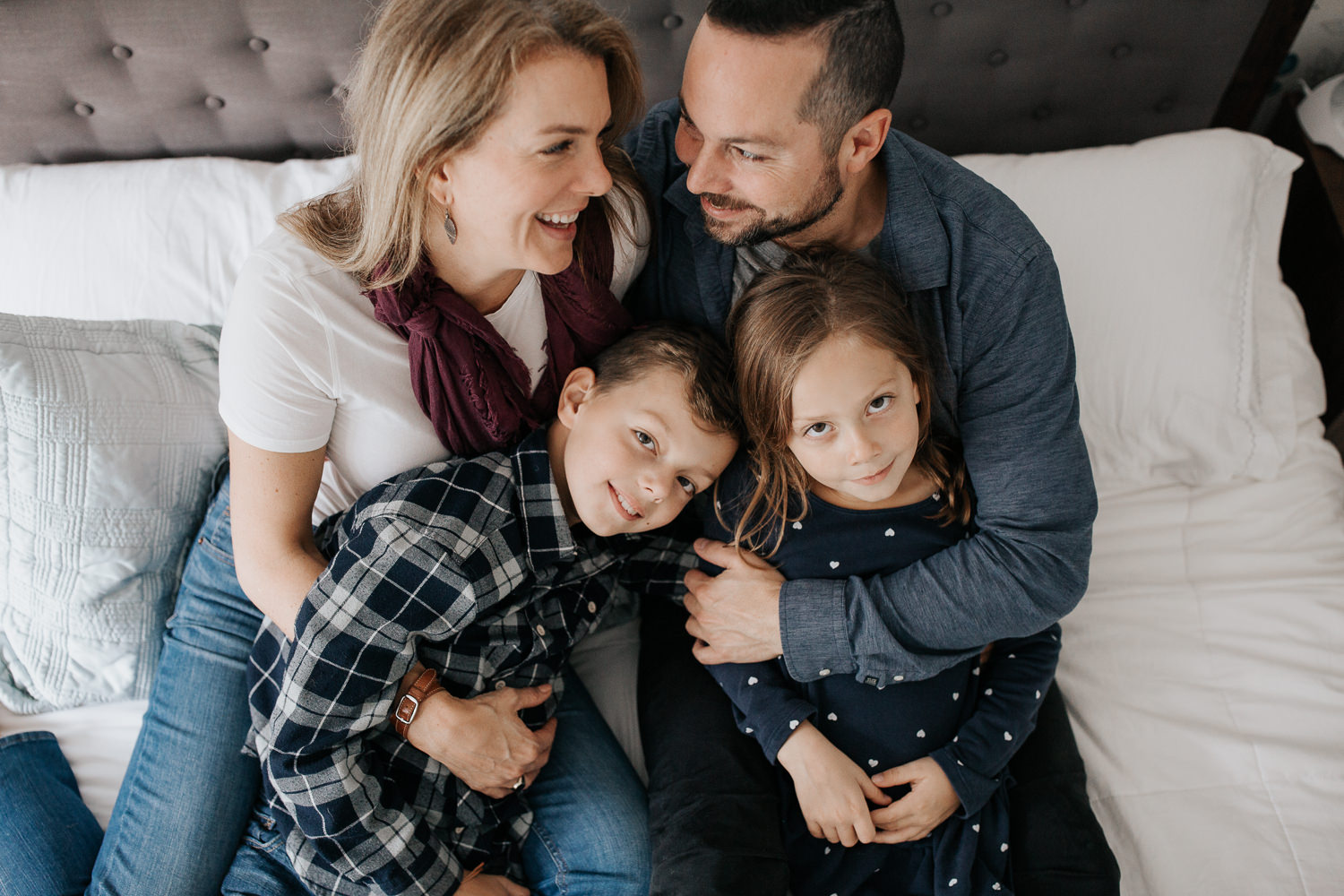 family of 4 sitting on master bed, 7 year old girl sitting in dad's lap, 9 year old boy in mom's, parents smiling at each other, kids looking up at camera - Markham Lifestyle Photography