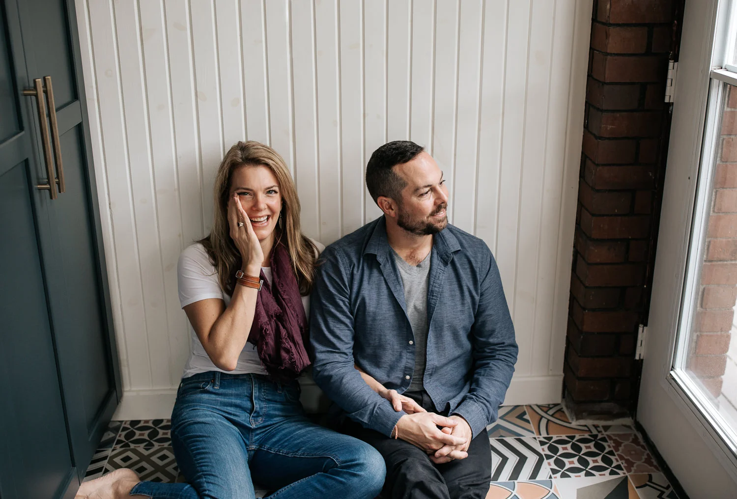 husband and wife sitting on ground of colourful tile, leaning against wall, arms linked, holding hands, man looking to side, woman's hand on cheek smiling at camera - York Region Lifestyle Photos