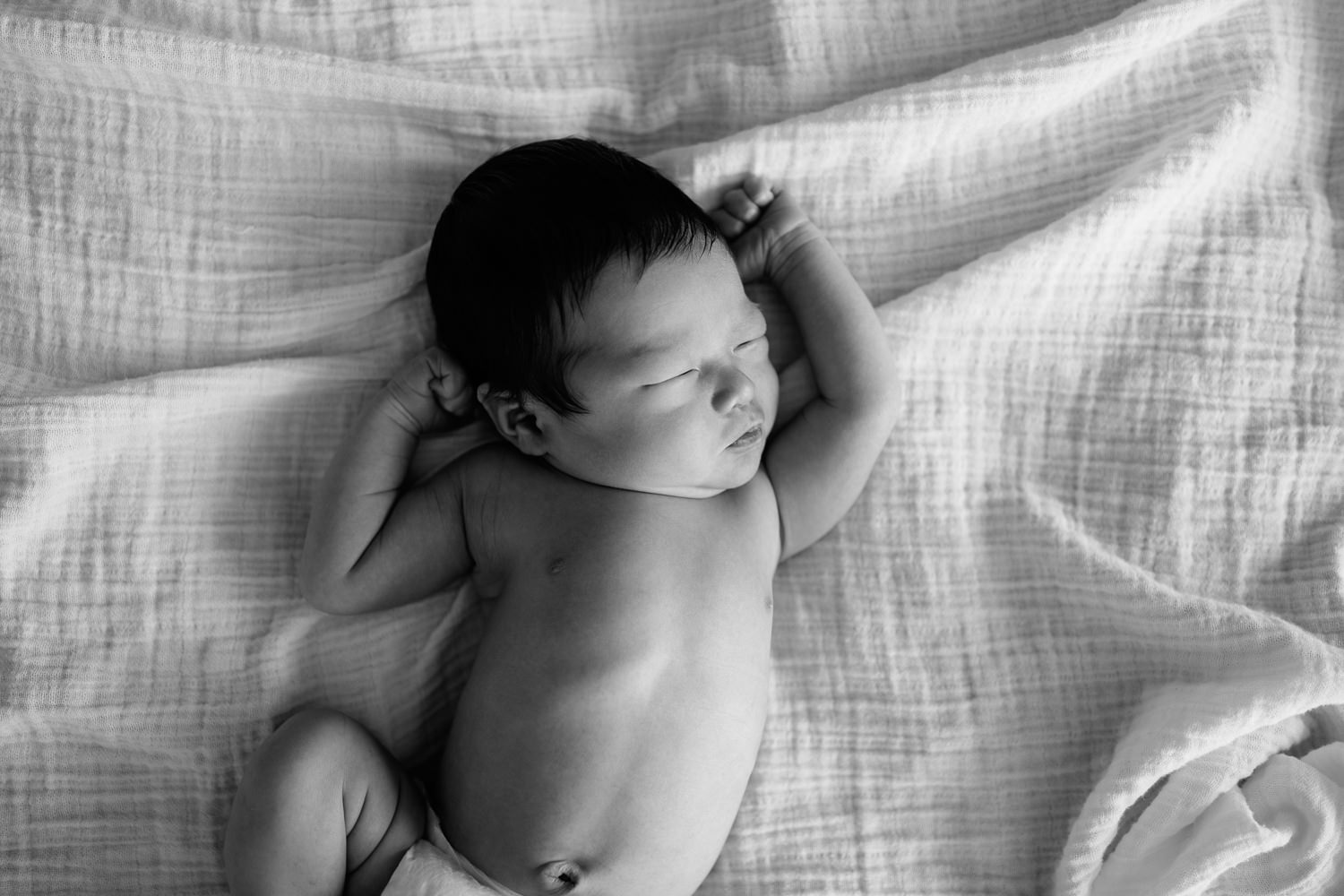 2 week old baby boy with long dark hair in diaper lying on bed, arms up by face, stretching, head to side, sleeping - Markham Lifestyle Photography