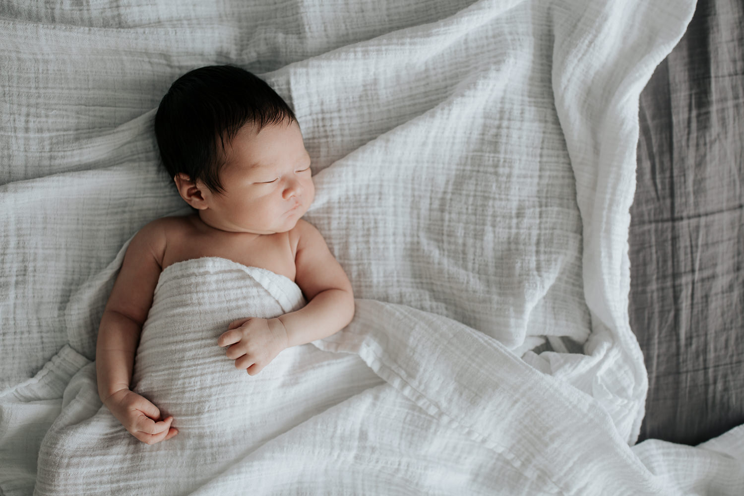 2 week old baby boy with long dark hair in diaper lying on bed, body covered by white swaddle, arms out, head to side, sleeping - Newmarket Lifestyle Photography